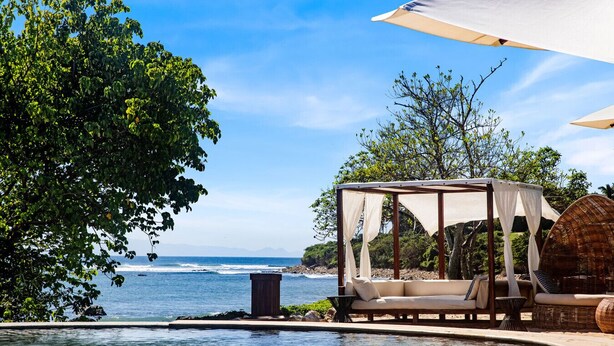 A poolside area with a canopy, a cushioned seating area, and a view of the ocean.