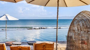 A beachside lounge area with a pool, wooden lounge chairs, and two umbrellas.
