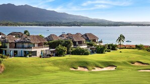 A golf course with a clubhouse, thatched-roof buildings, and a view of the water and mountains.