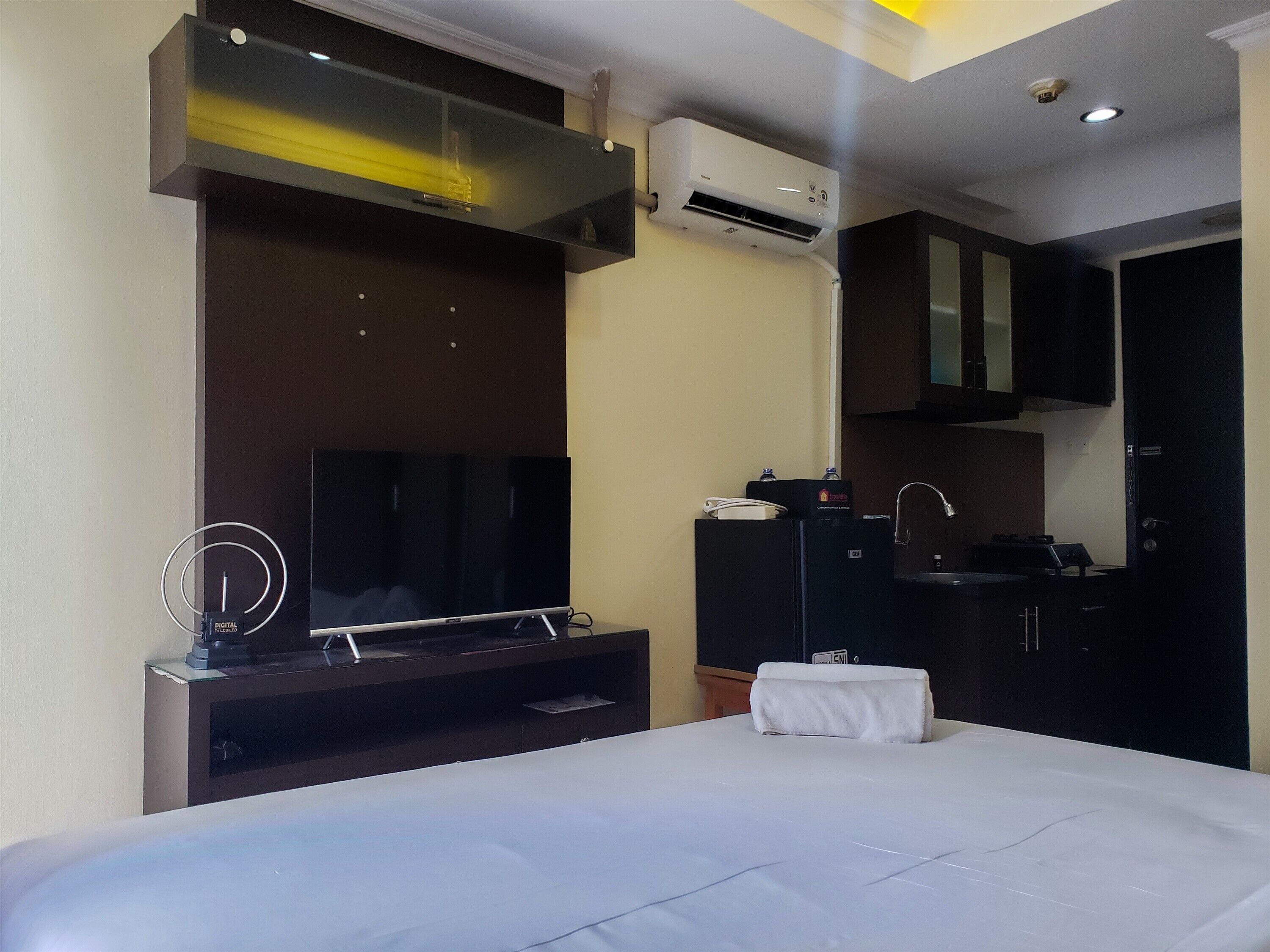 A hotel room with a bed, a TV, a small kitchenette, and an air conditioning unit.