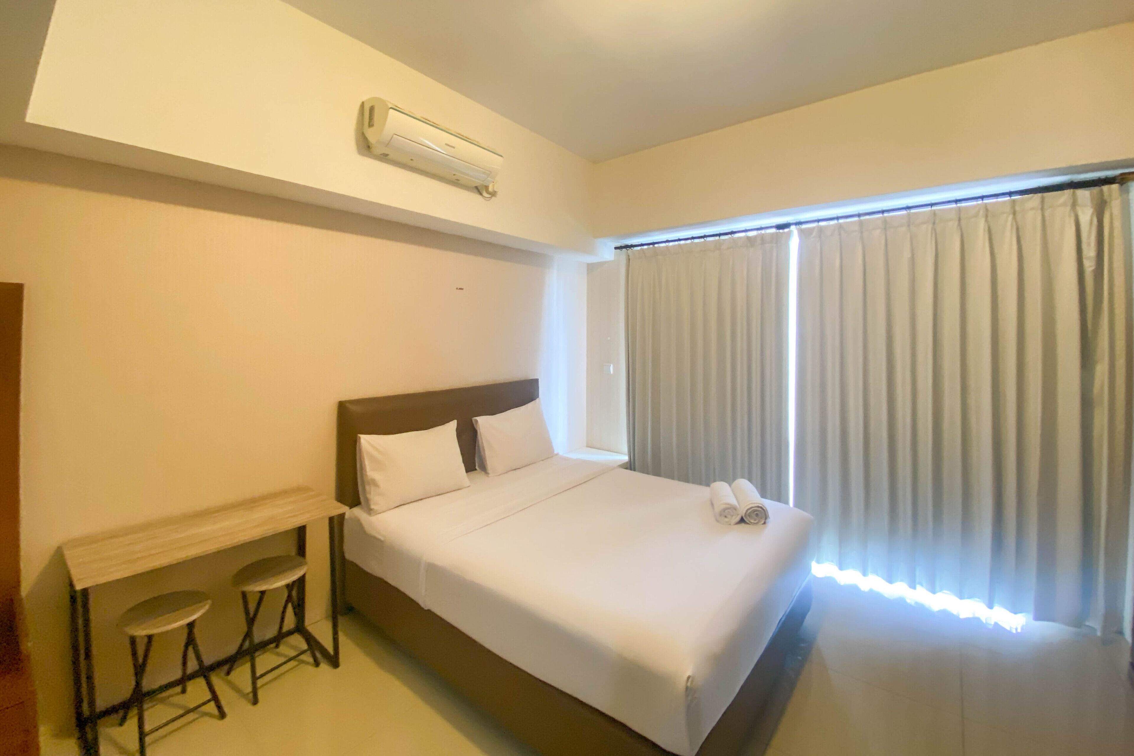 Una habitaci ón de hotel con cama, un escritorio y dos taburetes, una ventana con cortinas y un equipo de aire acondicionado.