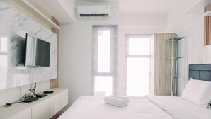 A modern hotel room with a large bed, a flat-screen TV mounted on a marble wall, a built-in air conditioning unit, and a glass shelving unit. - Simply Look Studio At Amazana Serpong Apartment (South Tangerang)