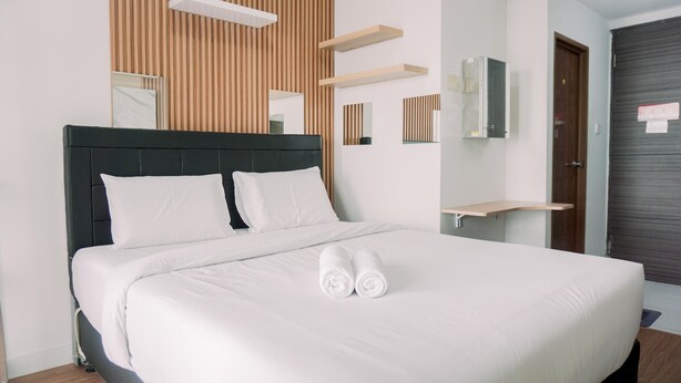 A neatly made bed with white linens and rolled towels, a wooden slat wall panel, and a mirror above the bed. - Simply Look Studio At Amazana Serpong Apartment (South Tangerang)