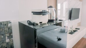 A modern hotel room with a bed, a coffee maker, a TV mounted on the wall, and a small desk with a laptop and remote controls. - Simply Look Studio At Amazana Serpong Apartment (South Tangerang)