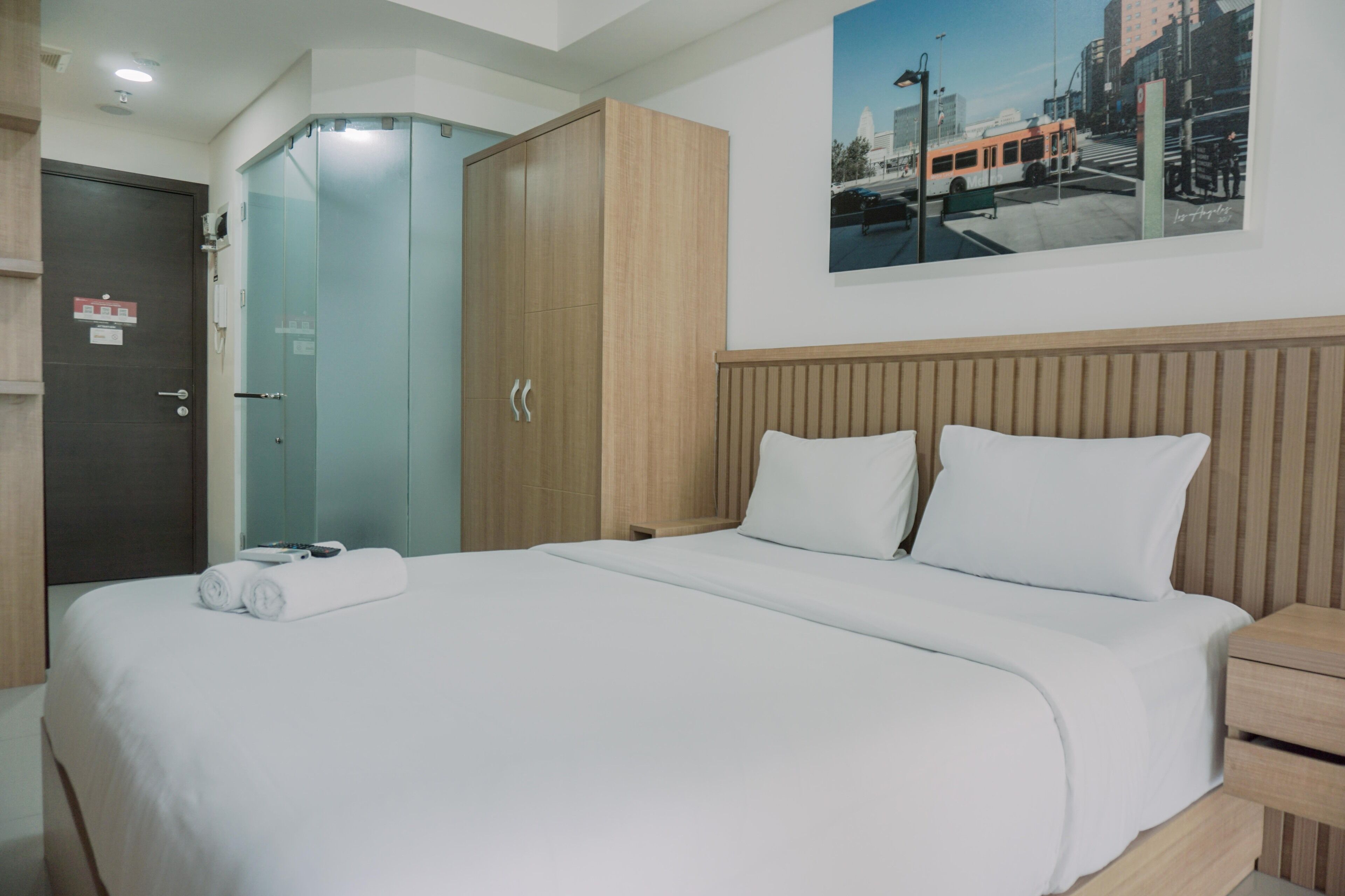 A hotel room with a large bed, a wooden wardrobe, a glass shower enclosure, and a cityscape painting on the wall.