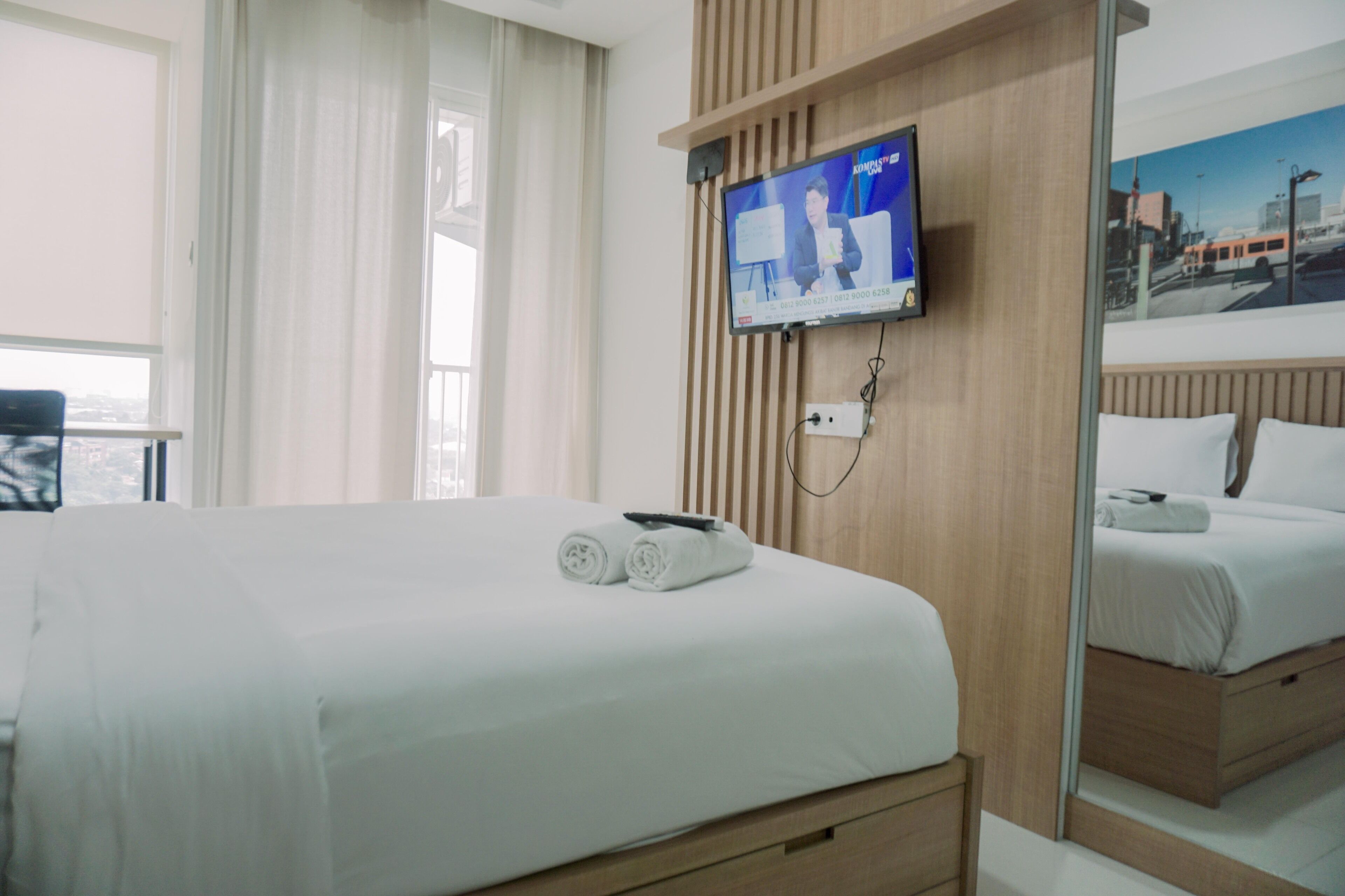 A hotel room with a large bed, a TV mounted on the wall, a window with curtains, and a mirror reflecting the bed.