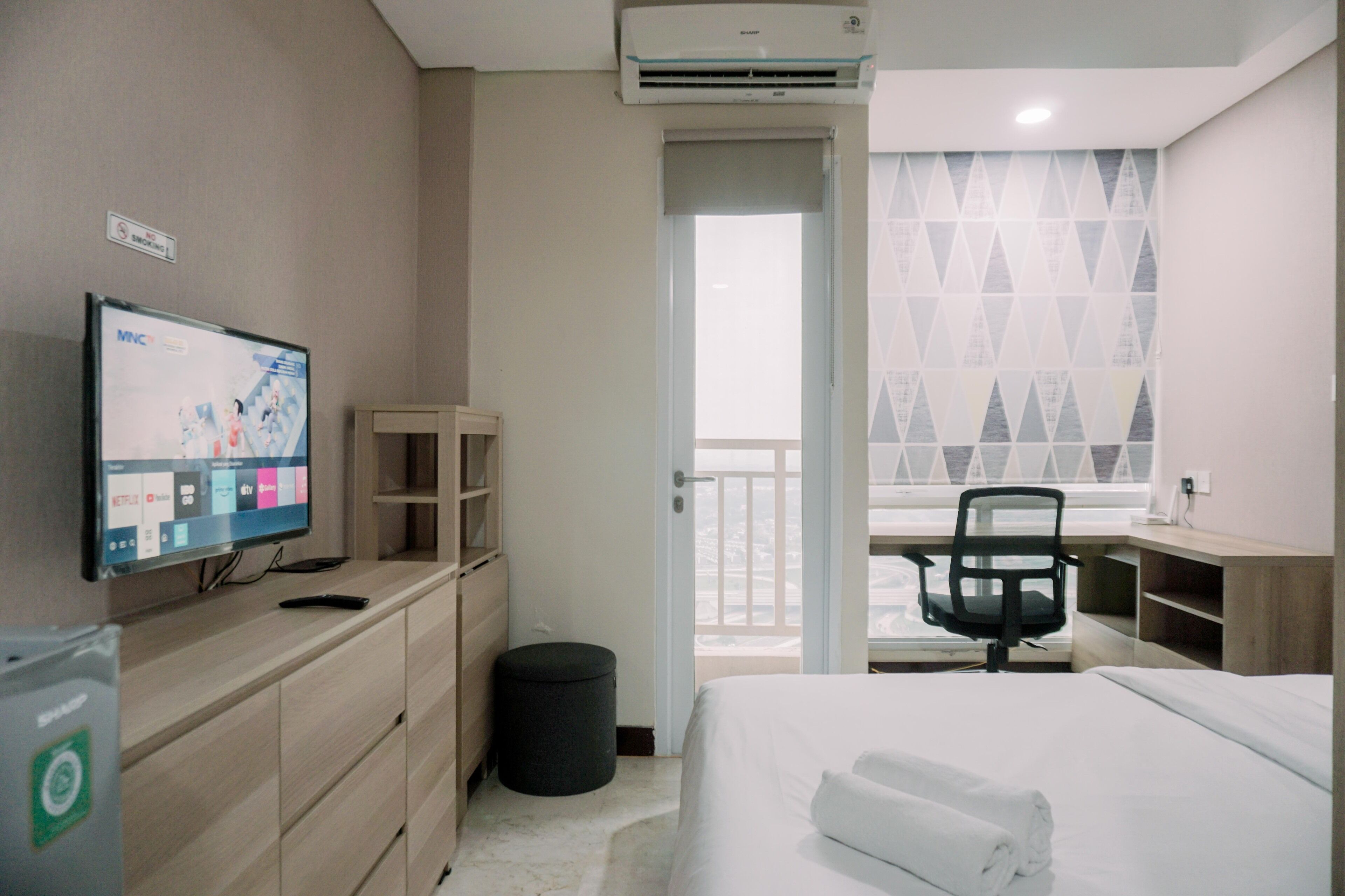 A hotel room with a bed, a desk, a chair, a TV, and a view of the outside.