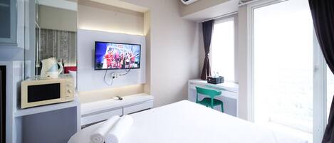 Habitación de hotel con cama, televisor montado en la pared, microondas, una pequeña mesa con silla y ventana con cortinas.