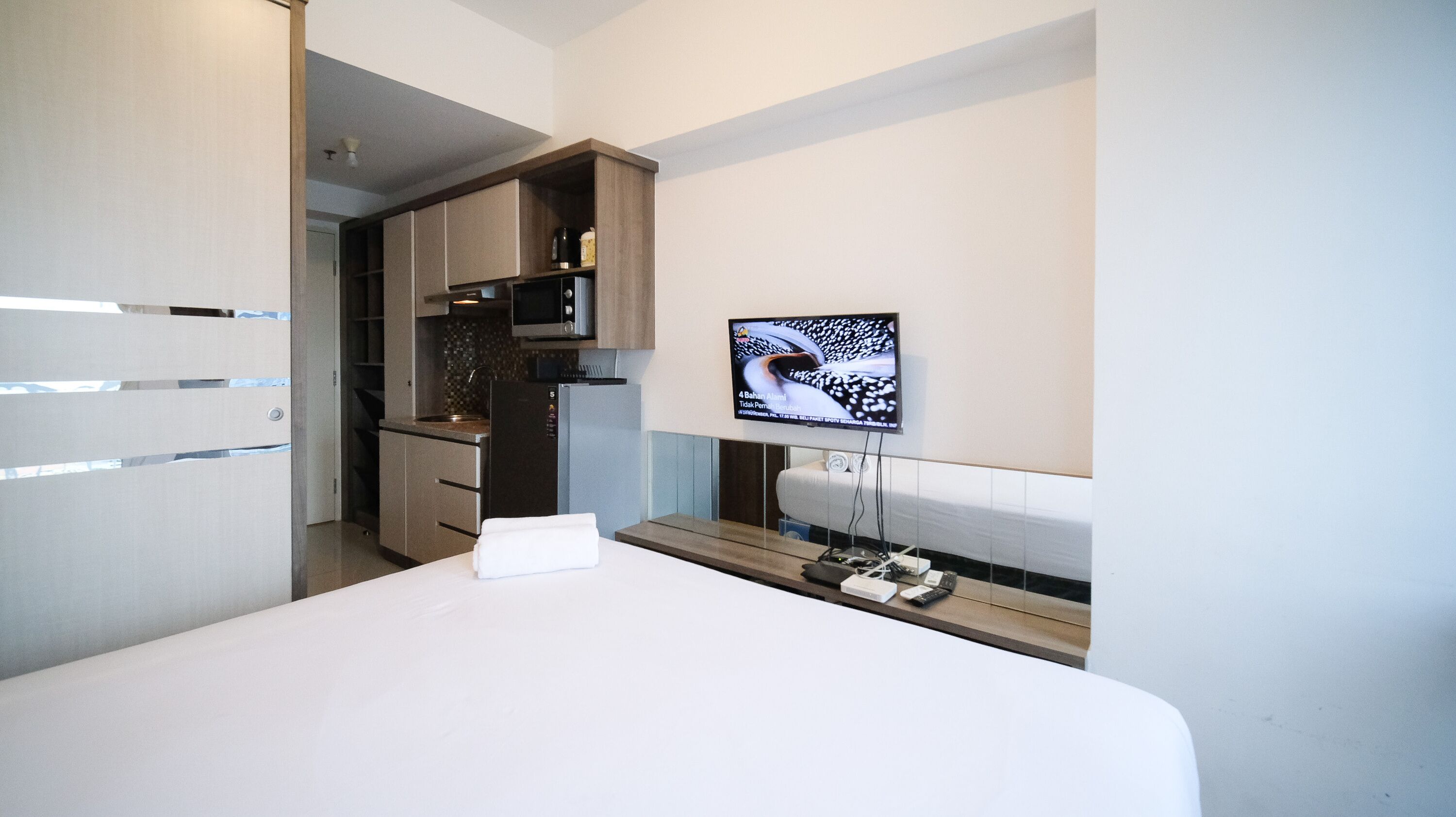 A modern hotel room with a bed, a TV mounted on the wall, a kitchen area, and a bathroom.