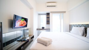 A hotel room with a bed, a TV mounted on the wall, a desk with a computer, and a window with a view. - Simple And Cozy Studio At Tanglin Supermall Mansion Apartment (Surabaya)