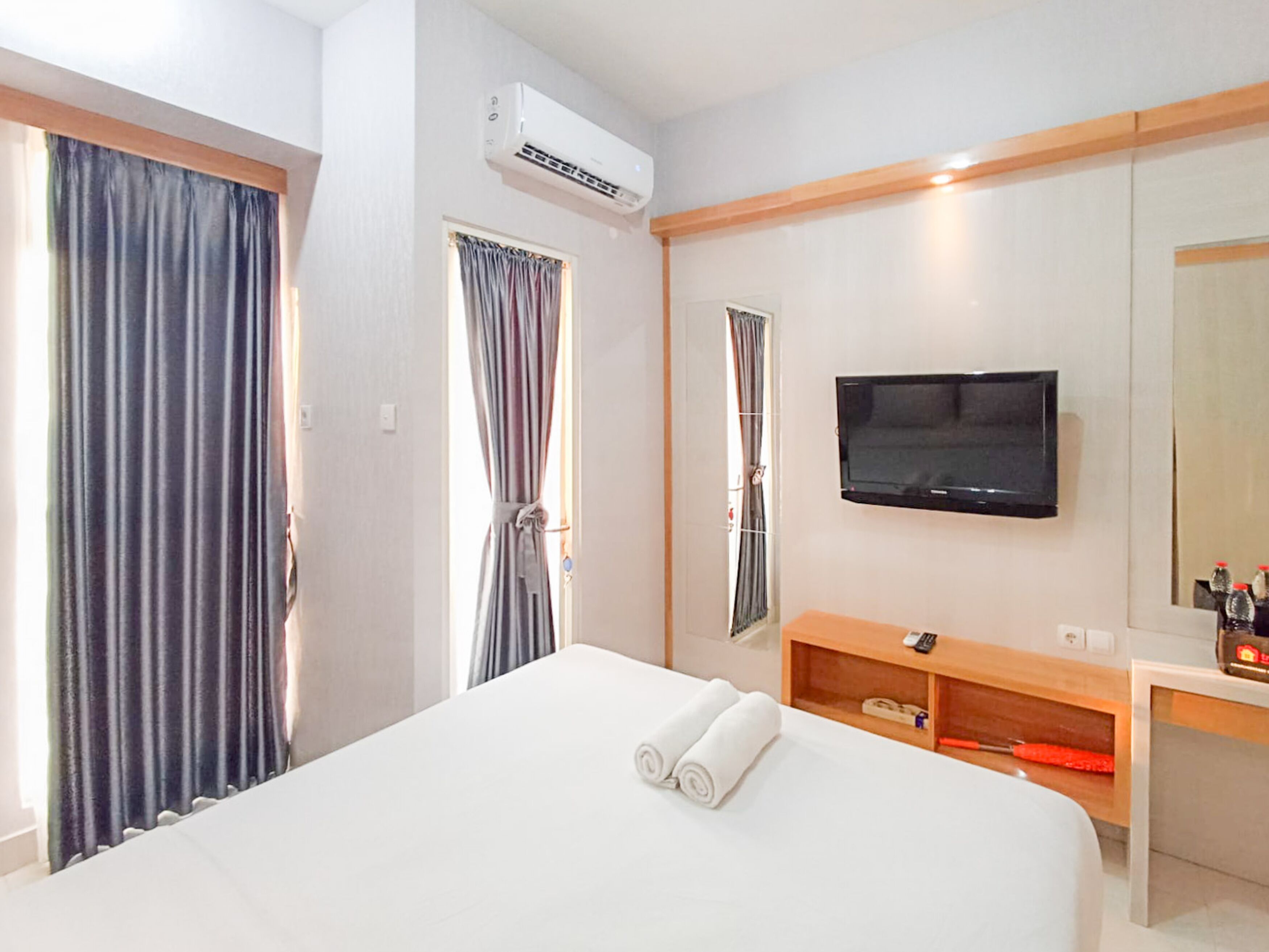 A hotel room with a bed, a TV mounted on the wall, a wooden nightstand, and a window with curtains.