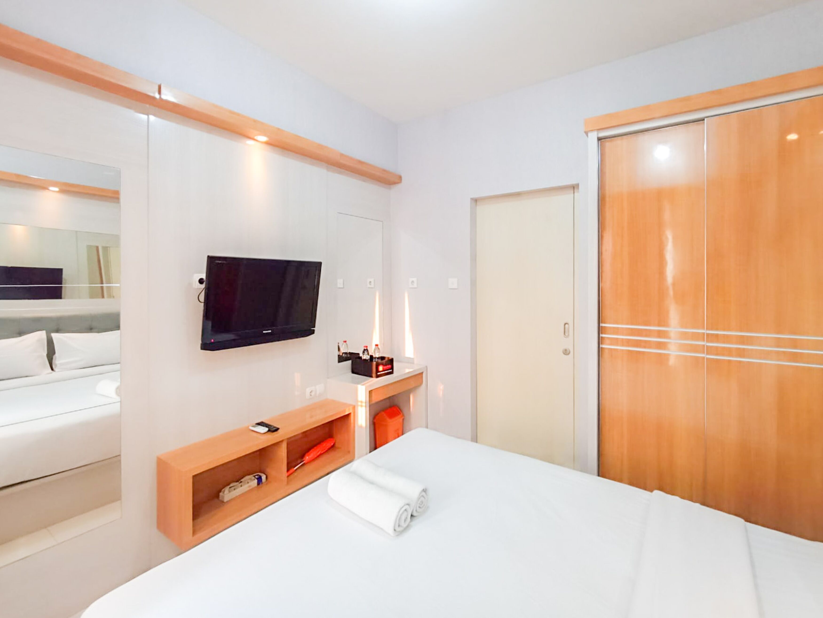 A modern hotel room with a bed, a TV mounted on the wall, a wooden wardrobe, and a small wooden shelf with a bottle and a remote control.
