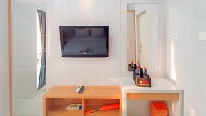 Habitación de hotel con televisor de pantalla plana montado en la pared, un escritorio de madera con control remoto, un estante con artículos de aseo y un espejo sobre el escritorio.