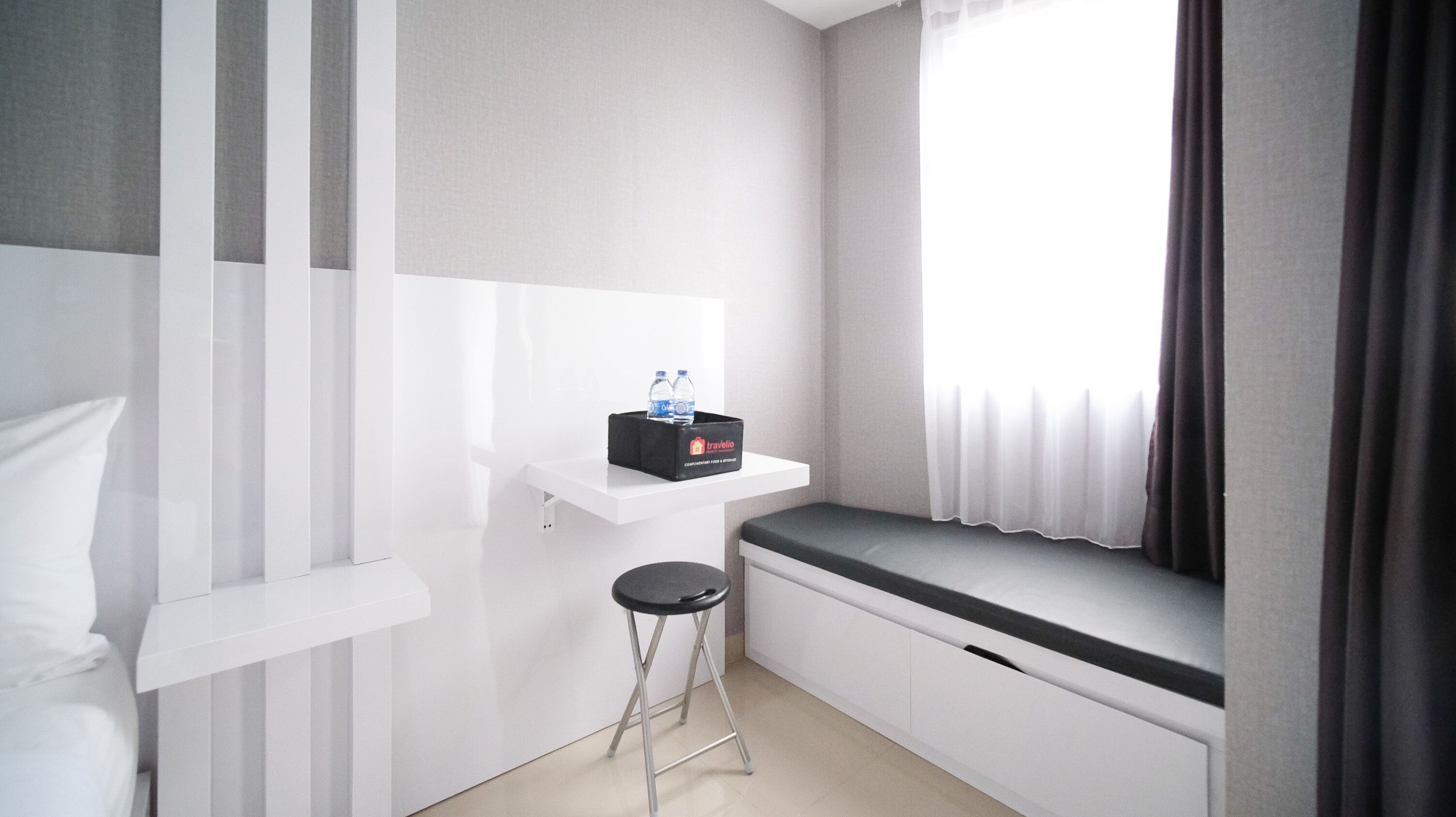 A small, modern room with a white bed, a white bench, a small stool, a white shelf with a water bottle, and a window with curtains.