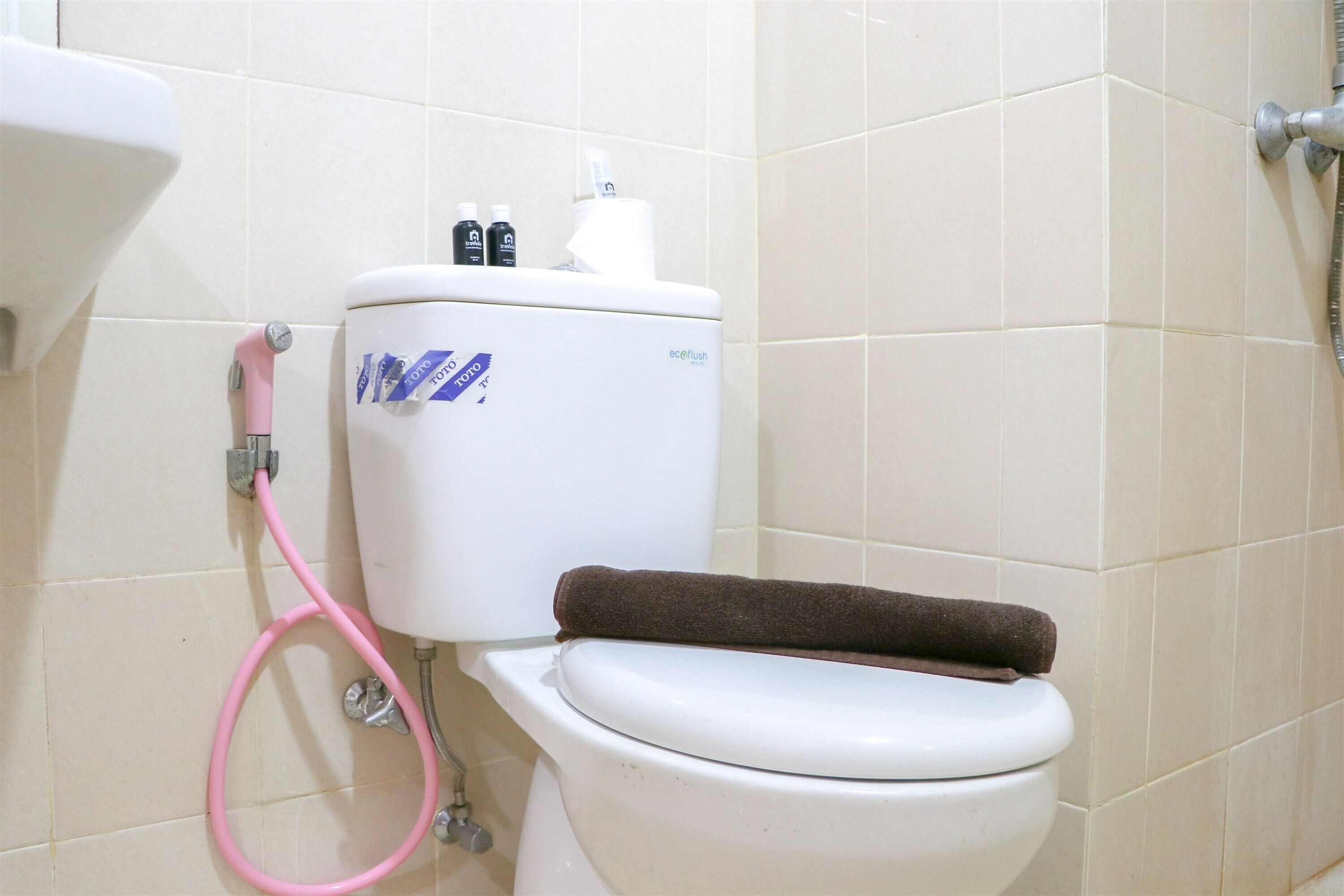 Inodoro con una manguera rosa, una toalla marrón y un porta papel higiénico con dos rollos.