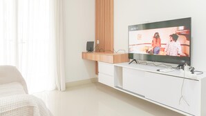 A modern living room with a flat-screen TV mounted on a white entertainment unit, a wooden accent wall, and a large window with sheer curtains. - Good Choice And Homey 2Br At Signature Park Grande Apartment (Jakarta)