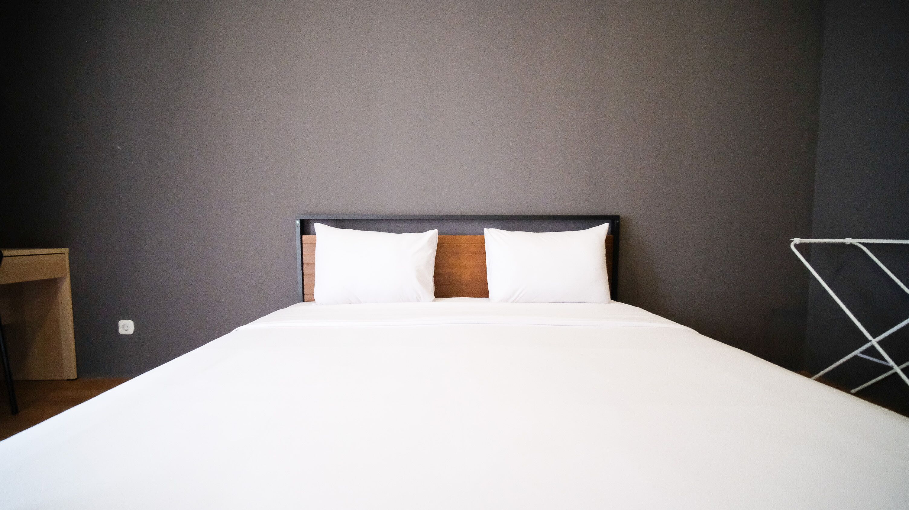 A neatly made bed with white linens and two pillows, a wooden headboard, and a grey wall.