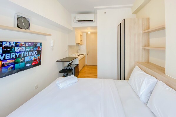 A hotel room with a bed, a TV mounted on the wall displaying various channels, a small desk with a chair, and a kitchenette area.
