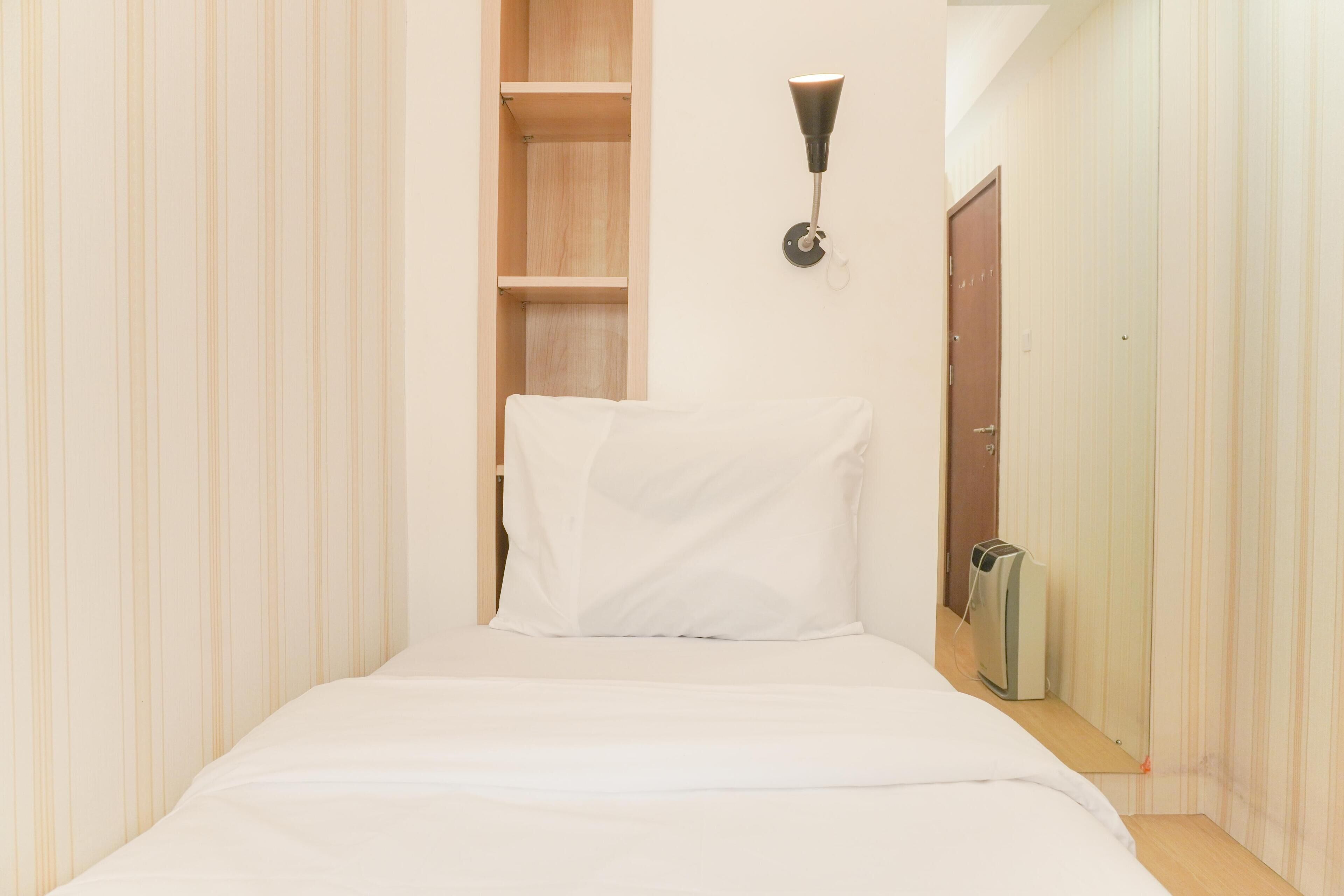 A narrow hotel room with a single bed, a wooden shelf, a wall-mounted light, and a door.