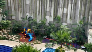 A children's play area with a slide and climbing structures, surrounded by lush greenery and a stone wall. - Nice And Homey 2Br Signature Park Grande Apartment (Jakarta)