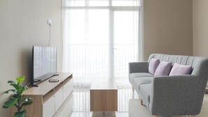 Ein modernes Wohnzimmer mit einem Flachbildfernseher auf einer Holzkonsole, einem grauen Sofa mit Kissen und einem großen Fenster mit transparenten Vorhängen.