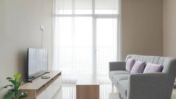 Una sala de estar moderna con un televisor de pantalla plana sobre una consola de madera, un sofá gris con cojines y un ventanal con cortinas translúcidas.