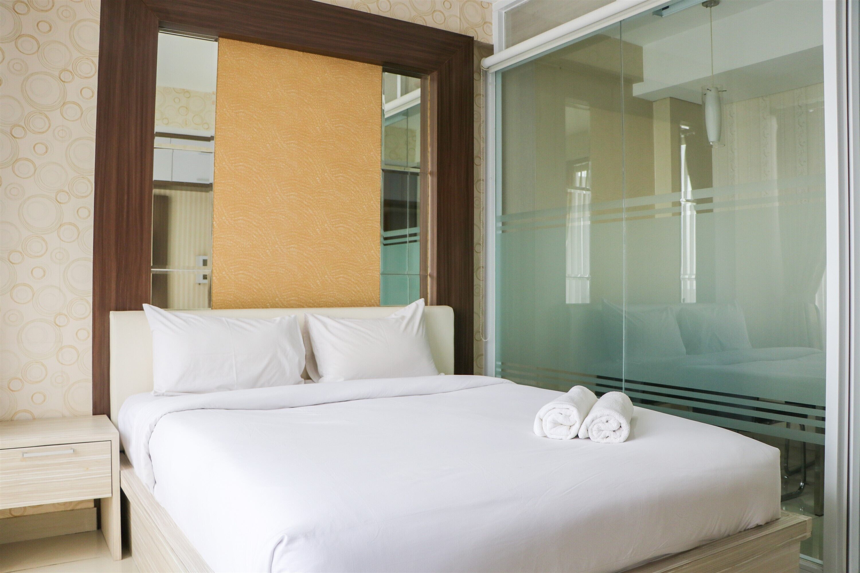 A neatly made bed with white linens and two rolled towels on the side, a wooden nightstand with a drawer, and a large mirror above the bed.