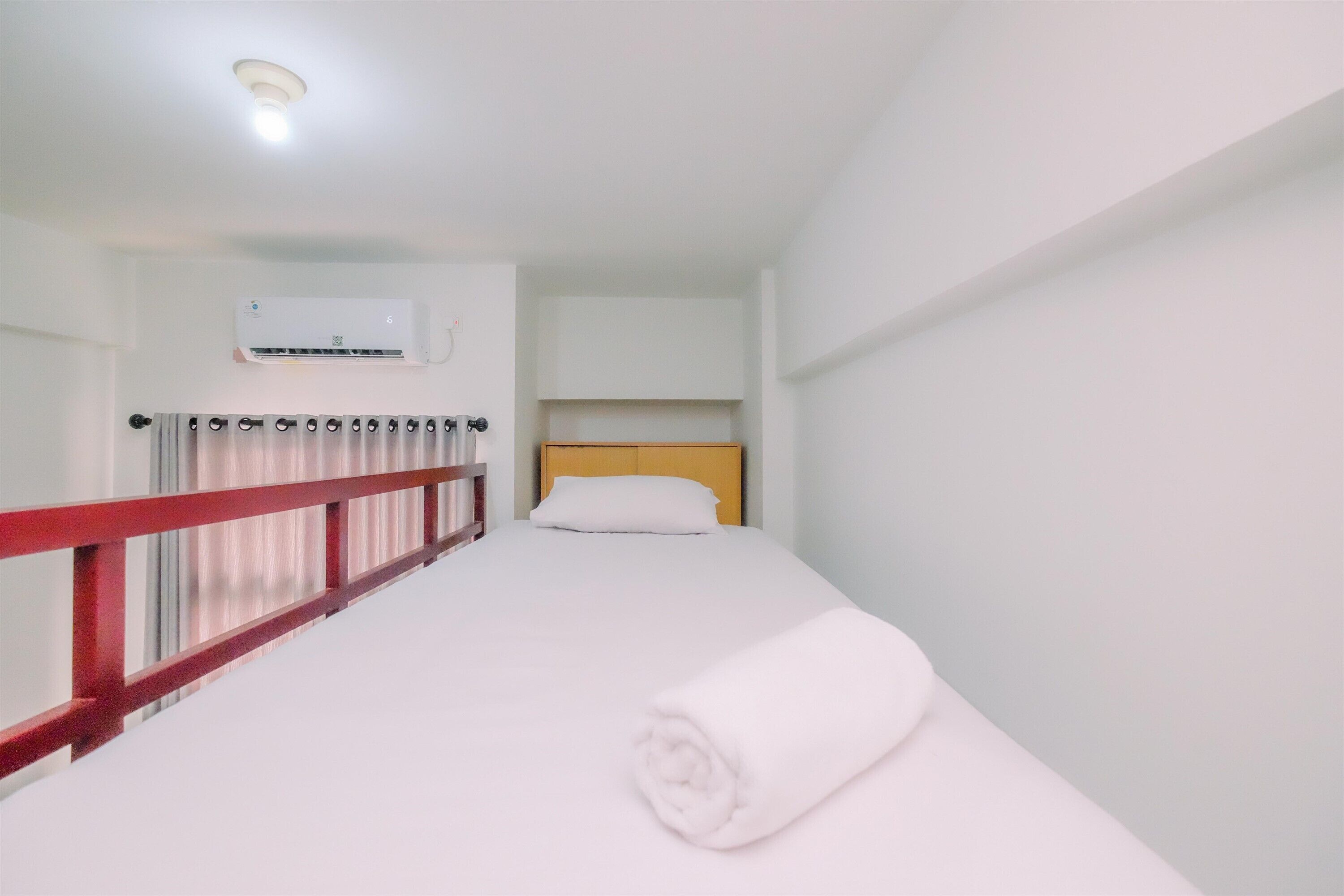 Un lit simple avec un matelas blanc et une serviette blanche enroulée, une tête de lit en bois, une barrière de sécurité rouge et un mur blanc sur lequel est fixé un appareil de climatisation.