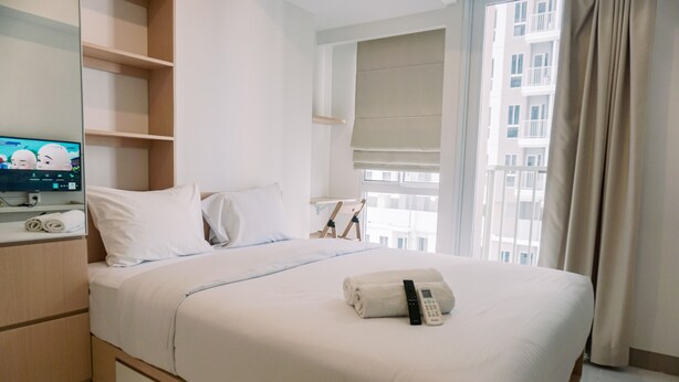 A neatly made bed with a remote control and a rolled towel, a television on a stand, and a window with a view of buildings. - Relax And Comfy Studio At Tokyo Riverside Pik 2 Apartment (Teluknaga)