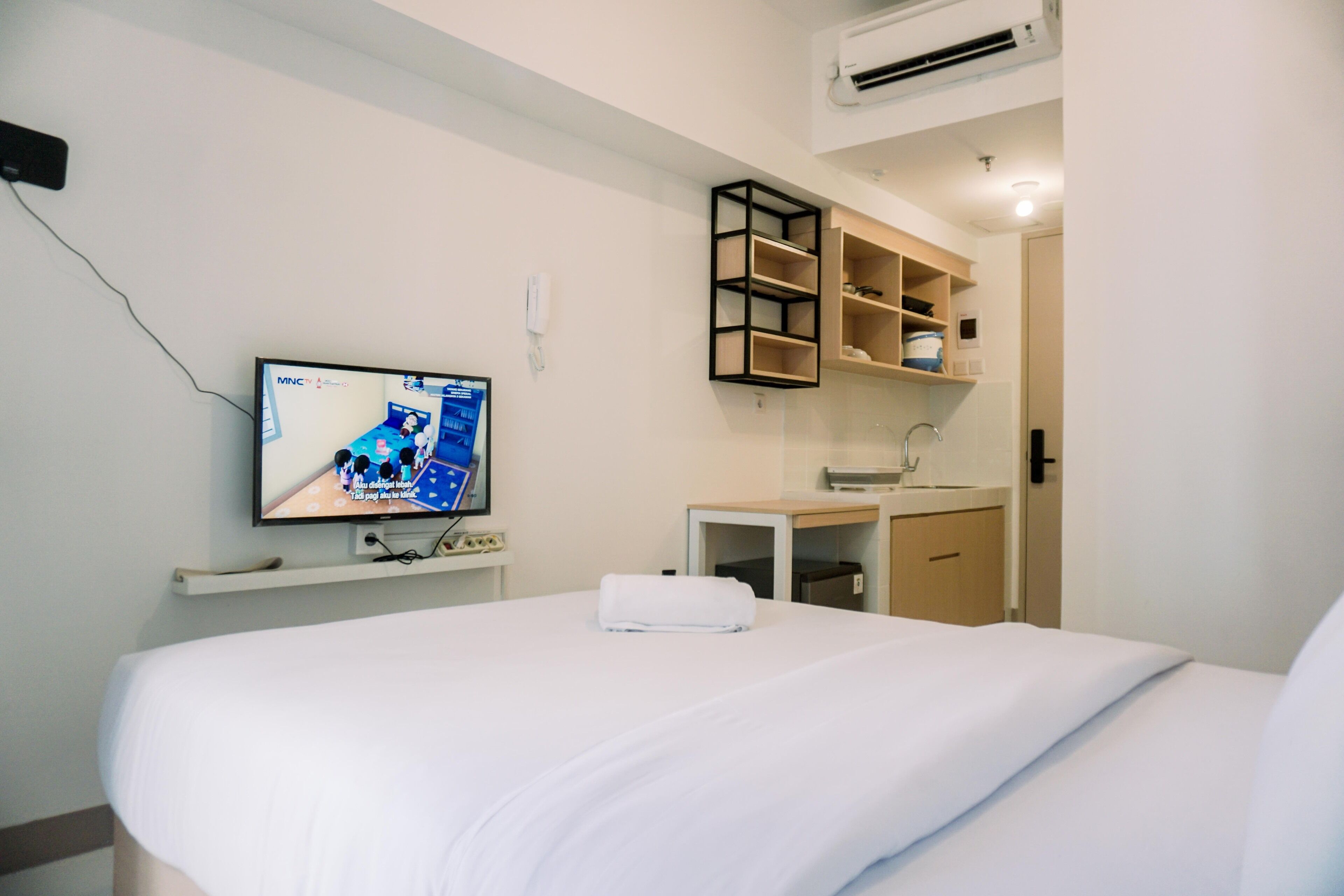 A hotel room with a bed, a TV mounted on the wall, a small desk with a sink, and a shelf unit.