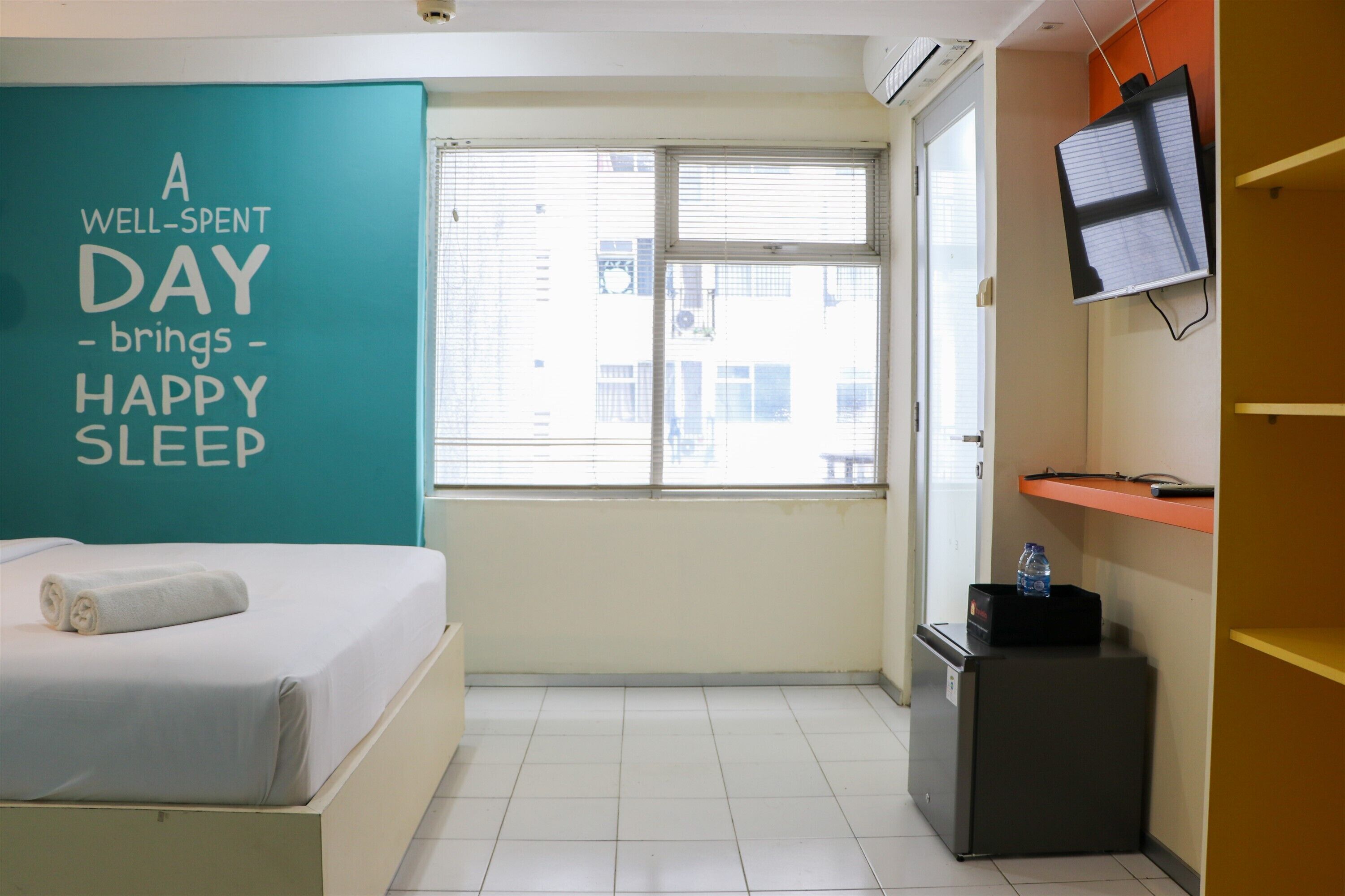 Ein Hotelzimmer mit Bett, einer türkisfarbenen Wand mit Motivationsspruch, einem Fenster mit Jalousien, einem an der Wand montierten Fernseher und einem kleinen Schreibtisch mit einer Wasserflasche.
