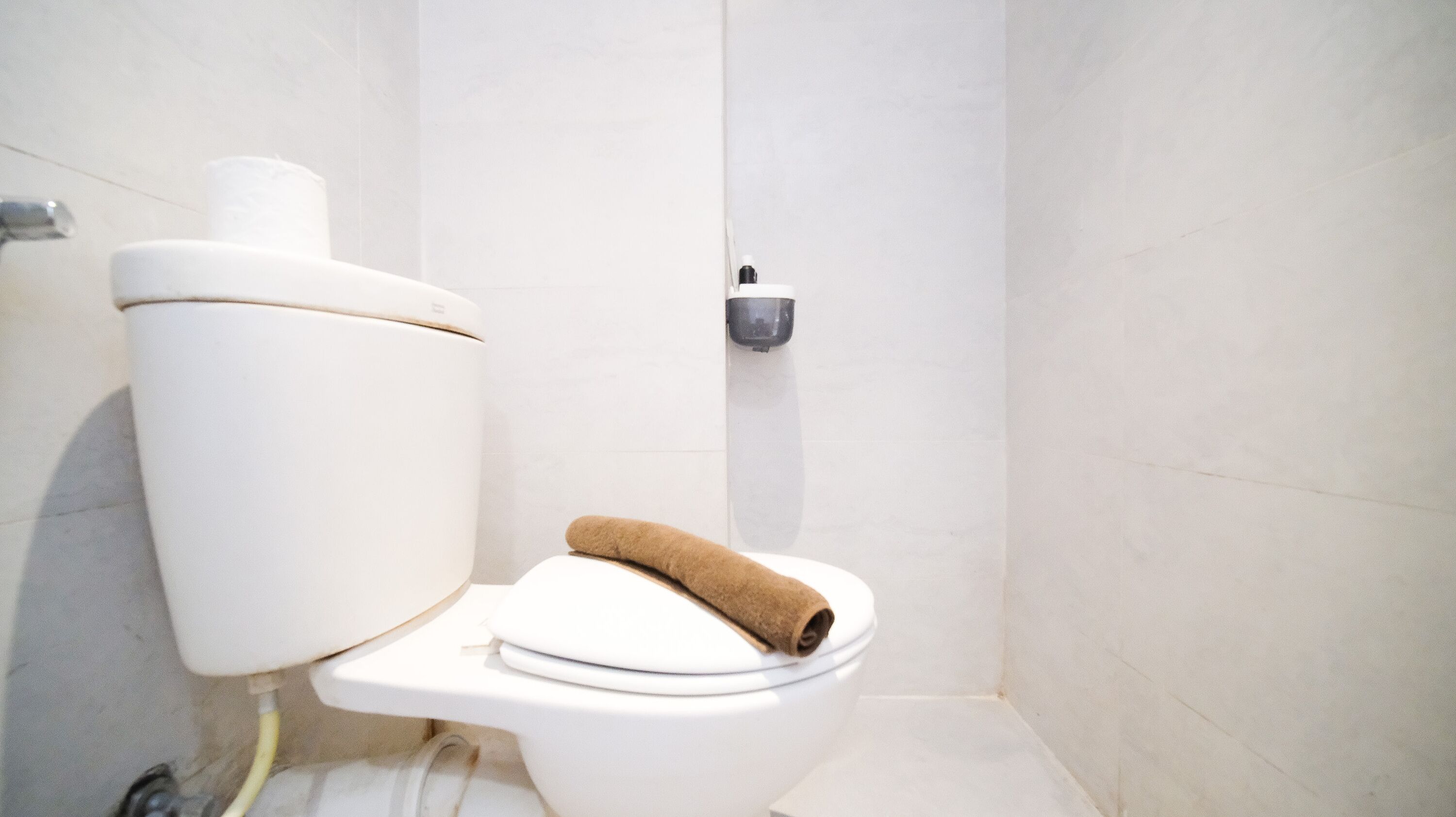 A white toilet with a brown towel on the seat, a toilet paper holder on the wall, and a visible plumbing pipe.