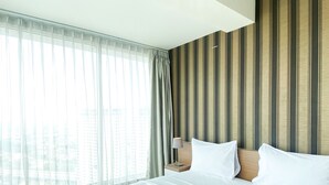 Ein Hotelzimmer mit einem großen Bett, einem Nachttisch, einer Lampe und Blick auf Gebäude durch transparente Vorhänge.