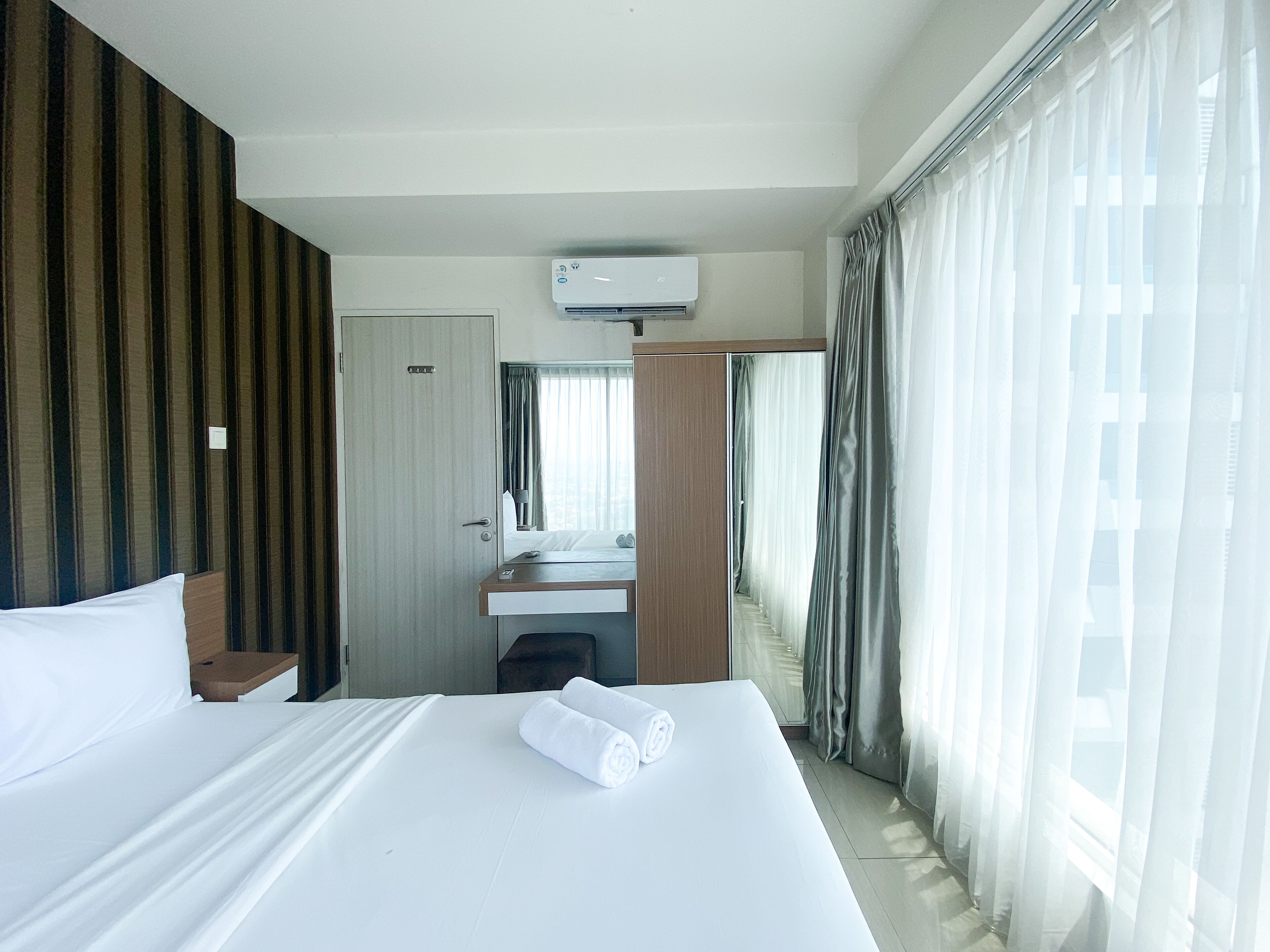 A hotel room with a large bed, a desk with a mirror, a window with curtains, and an air conditioning unit.