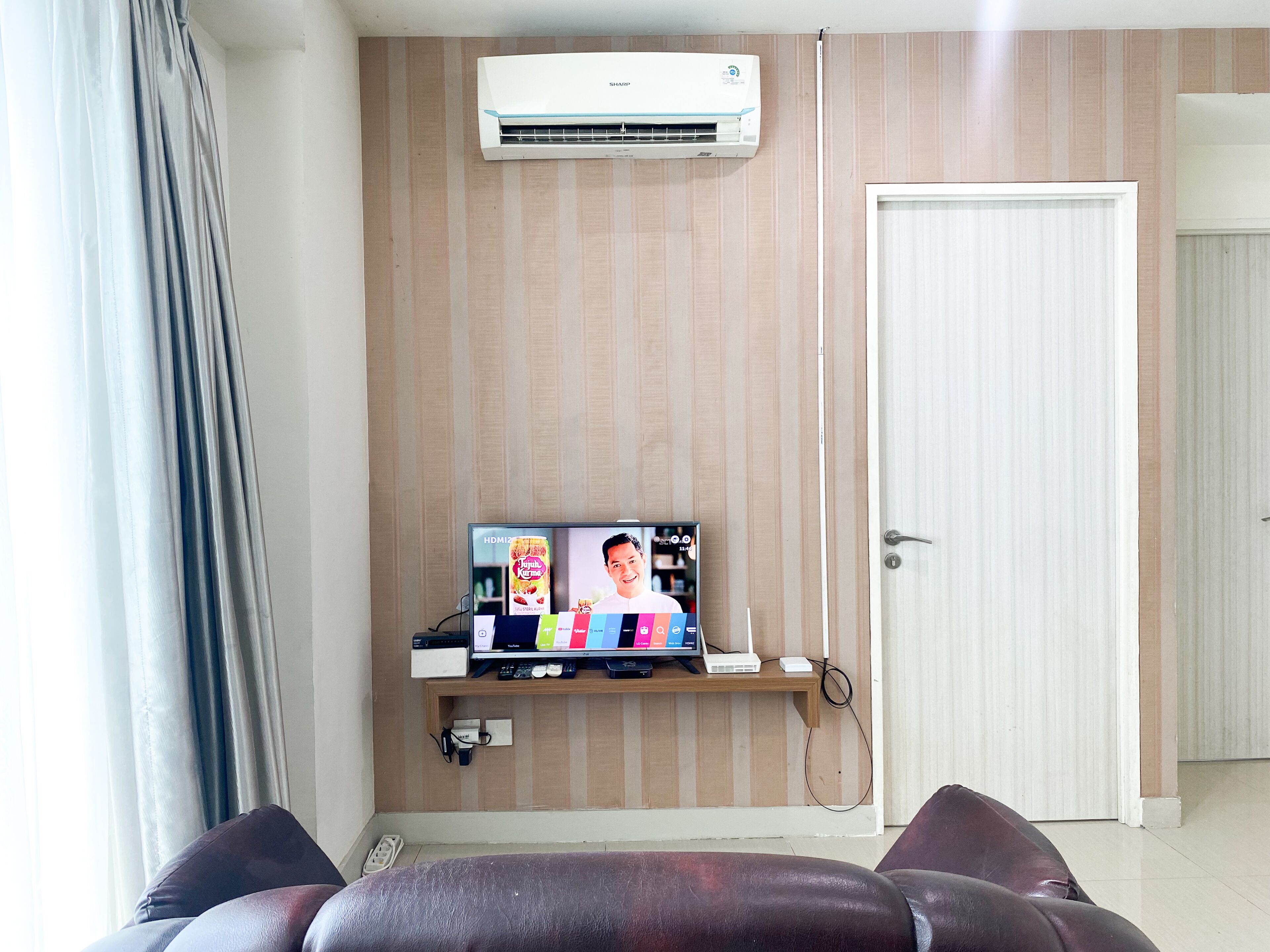 Quarto com ar condicionado fixado na parede, televisão sobre uma estante de madeira, porta e janela com cortinas.
