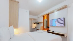 A modern hotel room with a bed, a TV mounted on the wall, a desk with a mirror, and a small kitchenette in the background. - Modern Look Studio At Urbantown Serpong Apartment (South Tangerang)