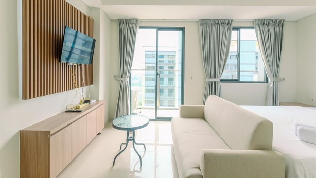 A hotel room with a bed, a sofa, a TV mounted on a wooden panel, a small table, and a large window with curtains. - Relaxing Studio At Daan Mogot City Apartment (Jakarta)
