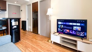 A modern living room with a flat-screen TV, a sofa, a coffee table, and a kitchen area with a refrigerator and a water dispenser. - Comfortable And Strategic 1Br At Vasanta Innopark Apartment (Cikarang)