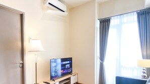 A hotel room with a TV on a wooden stand, a lamp, a sofa, and a window with curtains. - Comfortable And Strategic 1Br At Vasanta Innopark Apartment (Cikarang)