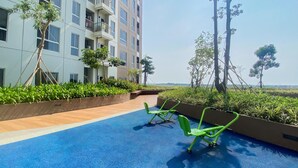 Uma área de piscina no terraço com piso azul, cadeiras de exercícios verdes e vista para um edifício moderno com sacadas.