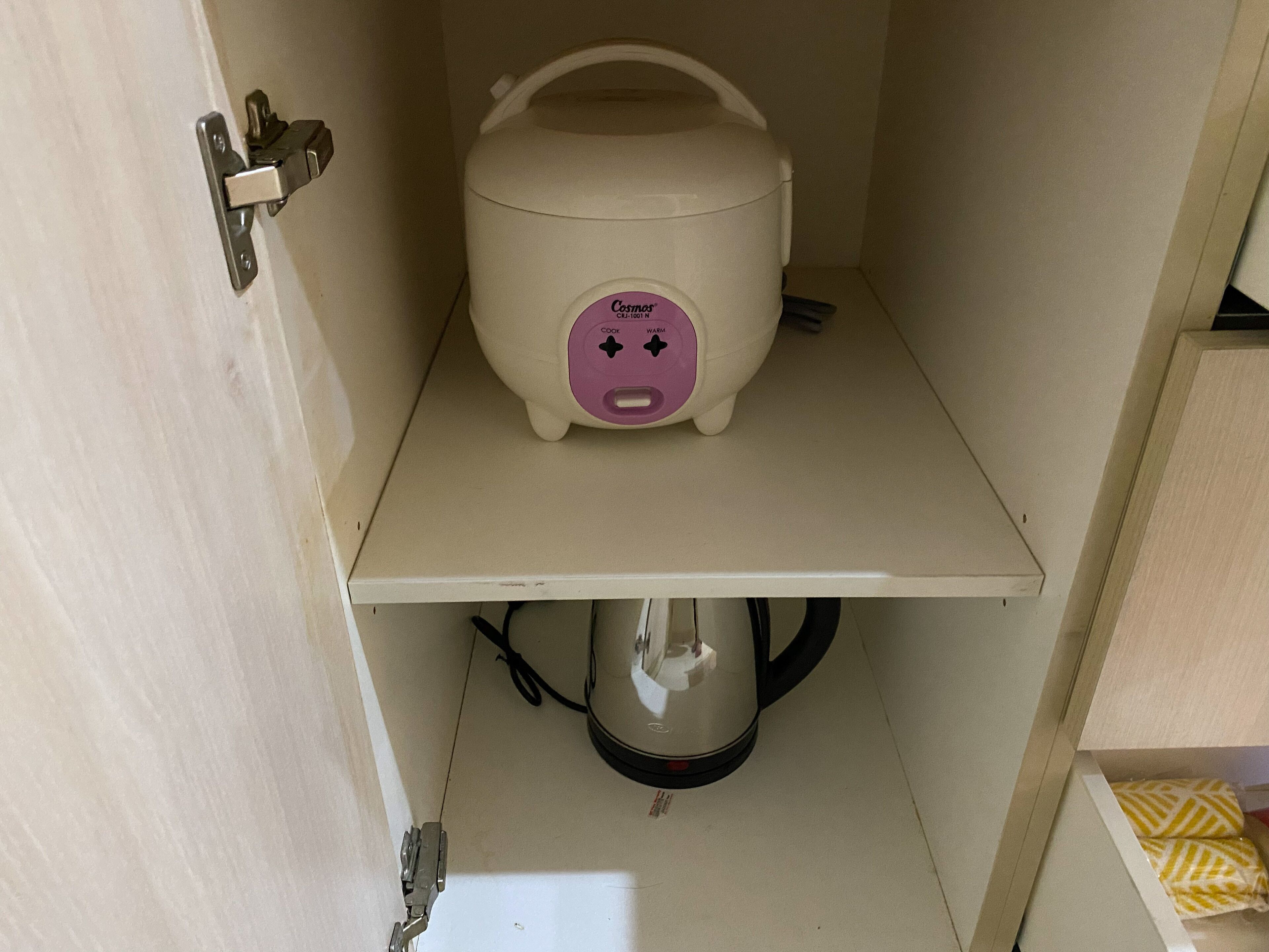 A compact kitchenette with a white countertop, a white electric kettle, and a small white appliance with a pink label.