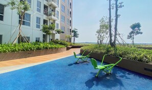 A rooftop pool area with blue flooring, green exercise chairs, and a view of a modern building with balconies. - Modern Look Studio Apartment At Tokyo Riverside Pik 2 (Teluknaga)