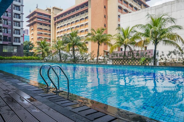 Uma piscina com deck de madeira, cercada por palmeiras e edifícios modernos.