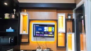 A modern living room with a flat-screen TV mounted on the wall, a coffee machine, and a small shelf with decorative items. - Brand New And Good Simply 1Br At Uttara The Icon Apartment (Depok)