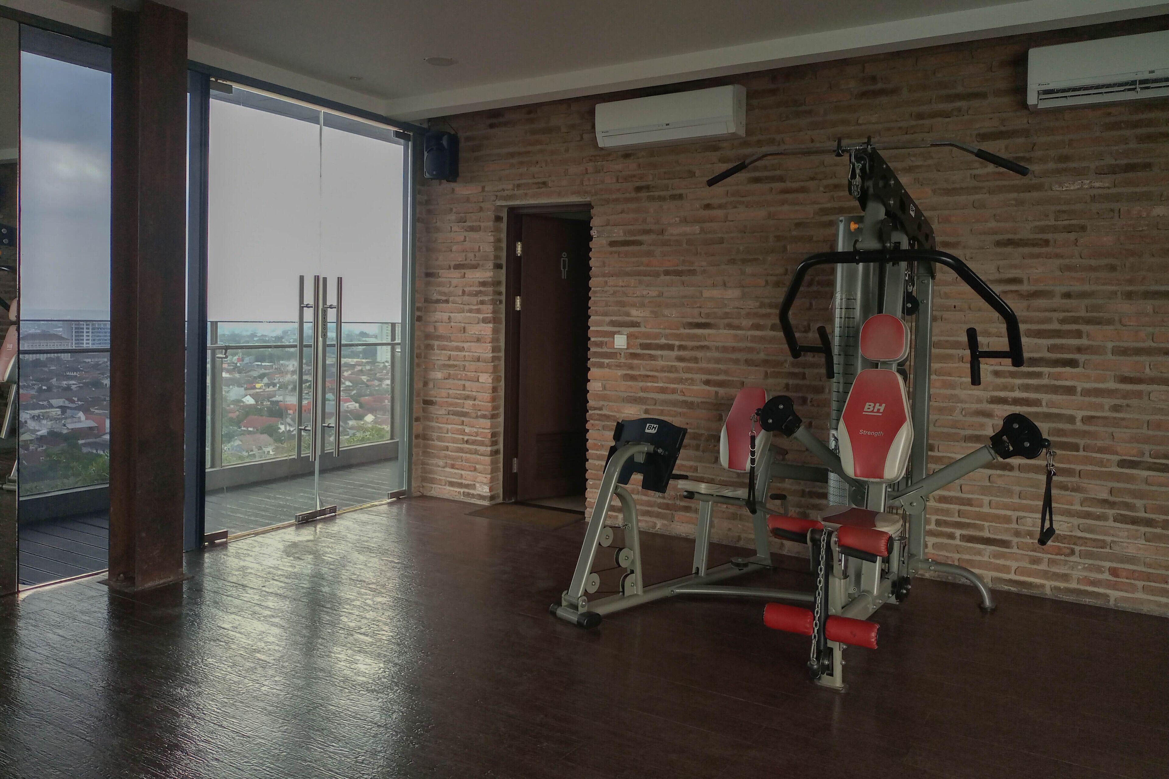 A spacious room with a large window offering a city view, a brick wall, and a ceiling-mounted air conditioning unit.