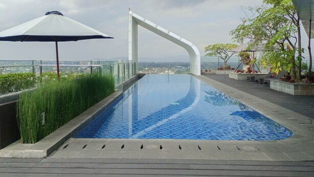 A swimming pool with a view of the city, surrounded by greenery and a modern architectural structure. - Brand New And Good Simply 1Br At Uttara The Icon Apartment (Depok)