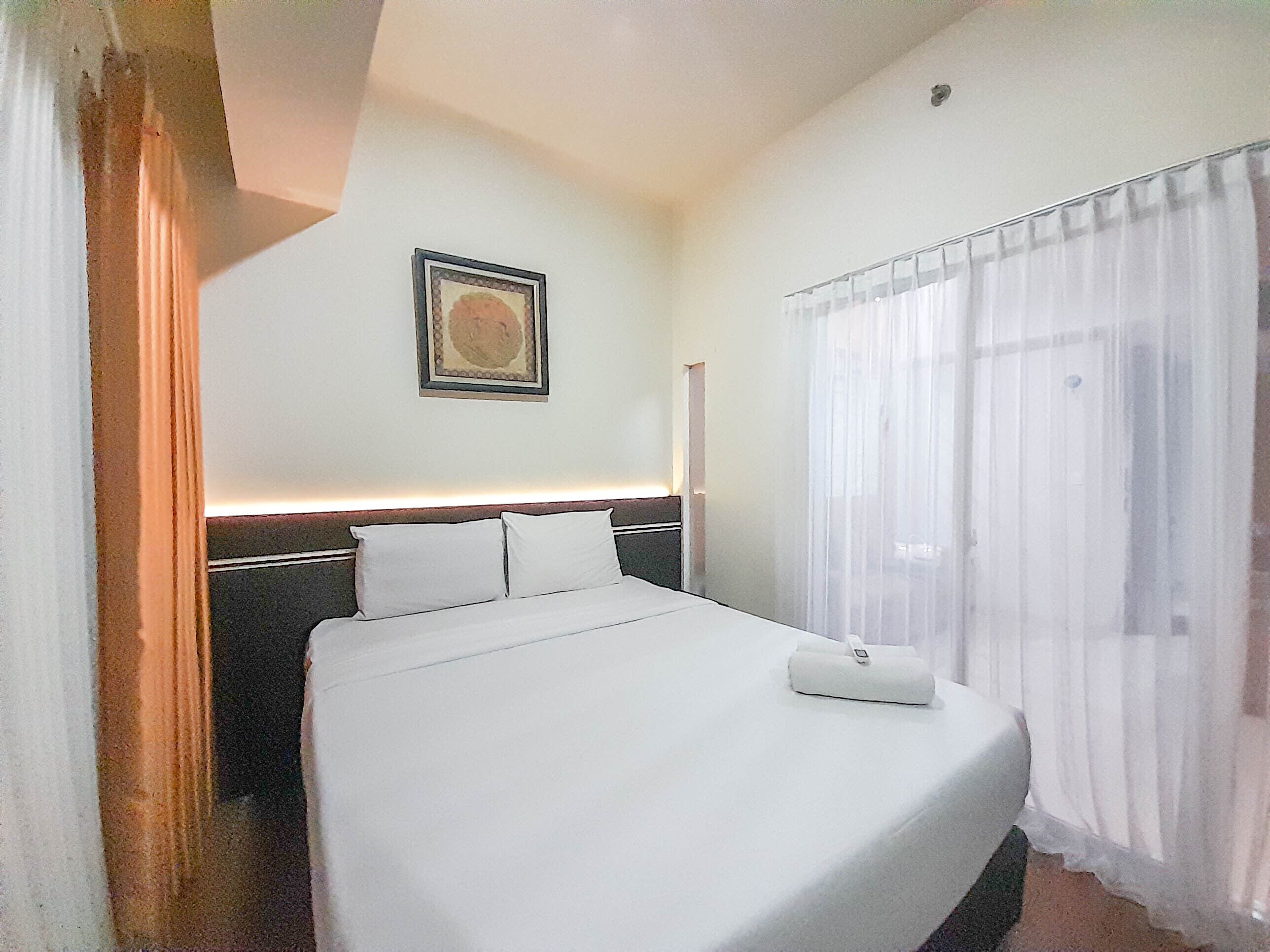 A hotel room with a neatly made bed, a framed picture on the wall, and sheer curtains on the window.