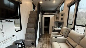 Een compact interieur met een bank, twee krukken, een aan de muur bevestigde televisie, een trap en een keukenhoek.