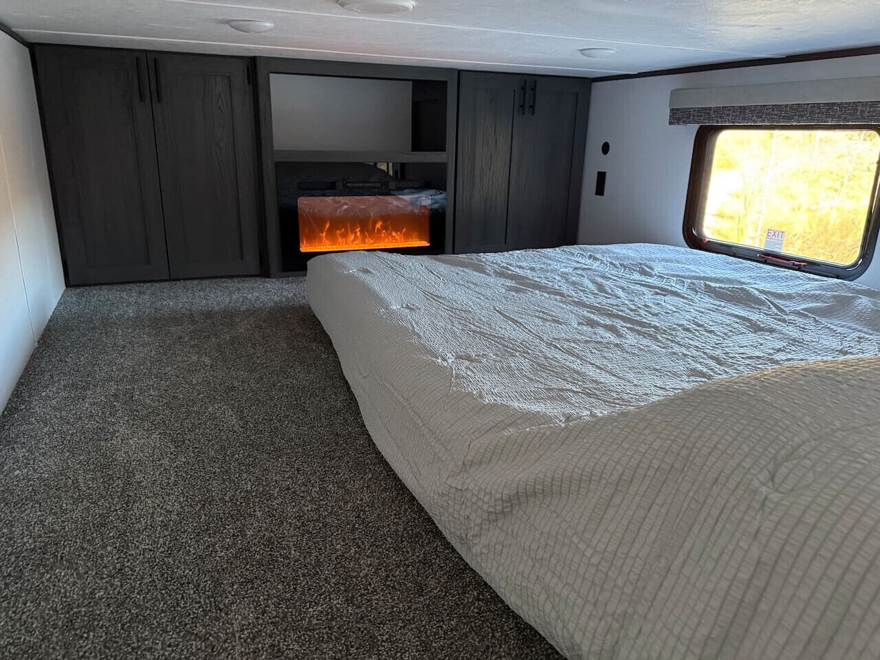 A compact RV bedroom with a bed, a small seating area, and a window providing a view of the outdoors.