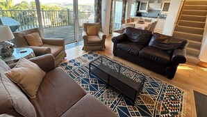 A spacious living area with a sofa, armchairs, and a coffee table. There is a balcony with a view of greenery and a kitchen in the background. - Firm Anchor (Salcombe)